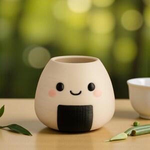 May include: A beige, onigiri-shaped planter with a smiling face, black eyes, and pink cheeks. A black nori seaweed detail is at the bottom. The planter is on a wooden surface with a blurred green background.
