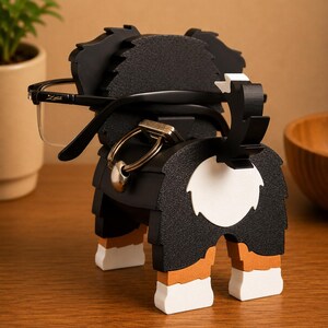 May include: A black, white, and brown dog-shaped eyeglasses holder. The holder has a textured surface and is holding a pair of black-framed glasses. The dog's tail is up, and the design includes a white circle on the rear.