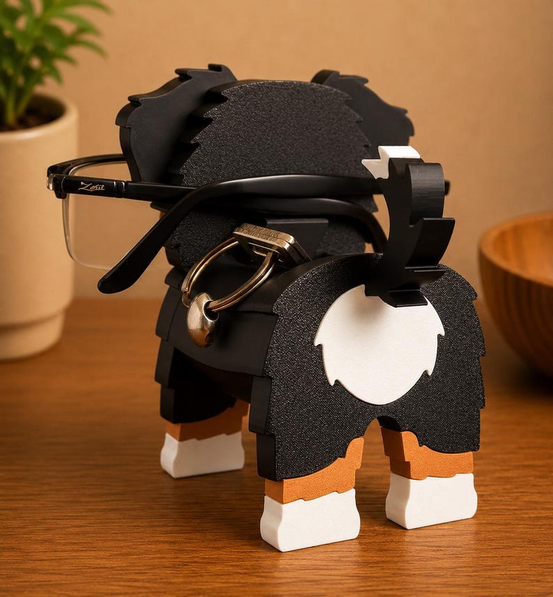 May include: A black, white, and brown dog-shaped eyeglasses holder. The holder has a textured surface and is holding a pair of black-framed glasses. The dog's tail is up, and the design includes a white circle on the rear.