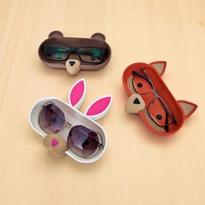 May include: Three whimsical eyeglass holders shaped like animals: a brown bear, a white bunny, and an orange fox. Each holder has a tray to hold glasses. The bunny holder has pink ears, and the fox has orange ears.
