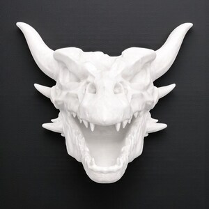 Puede incluir: Escultura de cabeza de drag&oacute;n blanca con boca abierta, dientes afilados y cuernos prominentes. La escultura est&aacute; sobre un fondo negro, resaltando la textura detallada y la forma de la criatura m&iacute;tica.