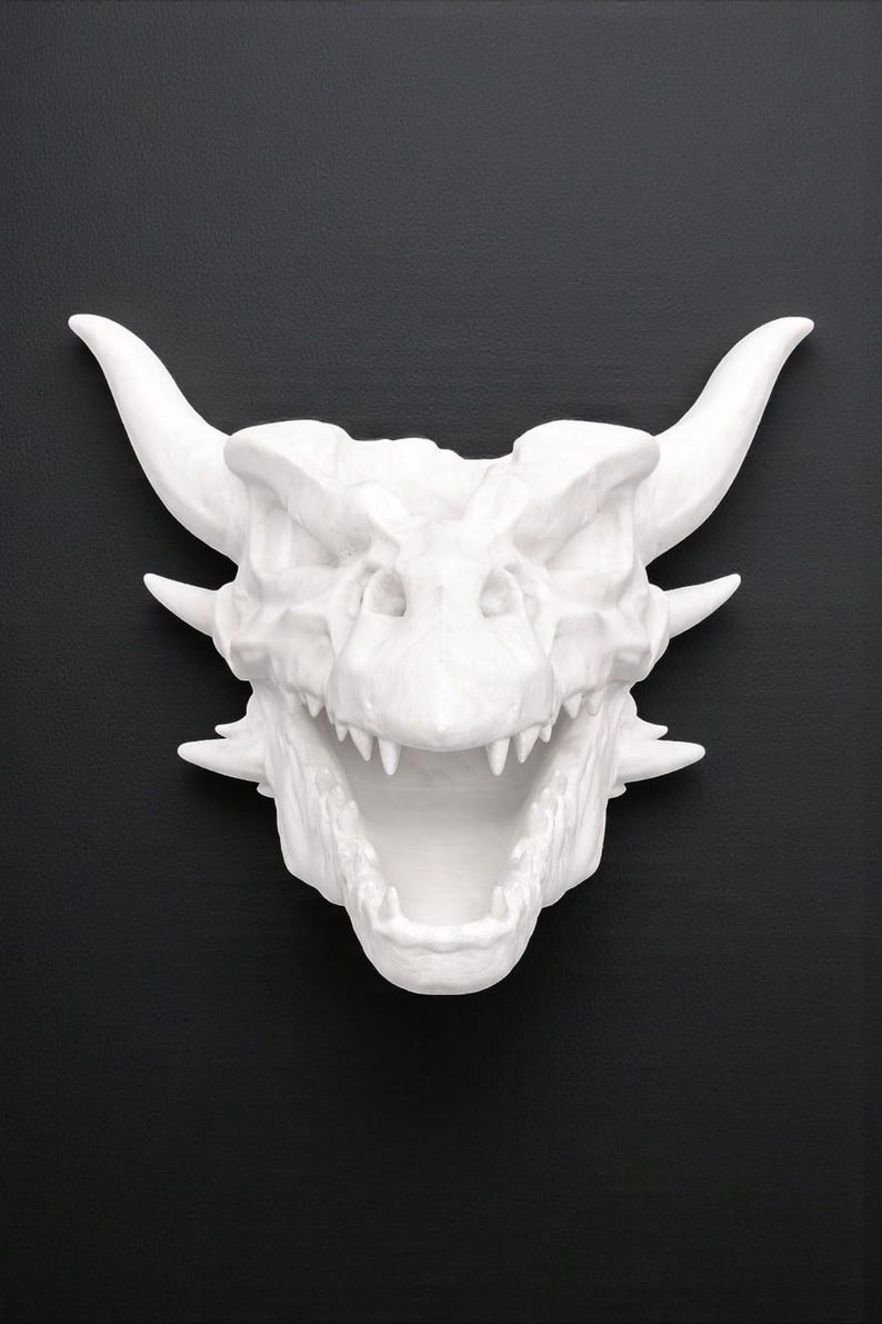 Puede incluir: Escultura de cabeza de drag&oacute;n blanca con boca abierta, dientes afilados y cuernos prominentes. La escultura est&aacute; sobre un fondo negro, resaltando la textura detallada y la forma de la criatura m&iacute;tica.