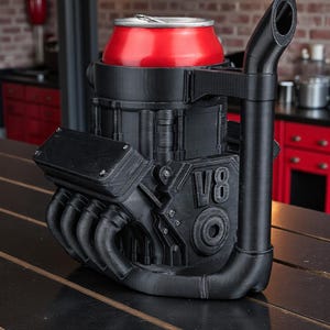 May include: A black V8 engine-shaped can cooler holds a red can. The cooler has detailed mechanical features, including exhaust pipes and the letters "V8". It sits on a dark wooden surface, with a brick wall in the background.