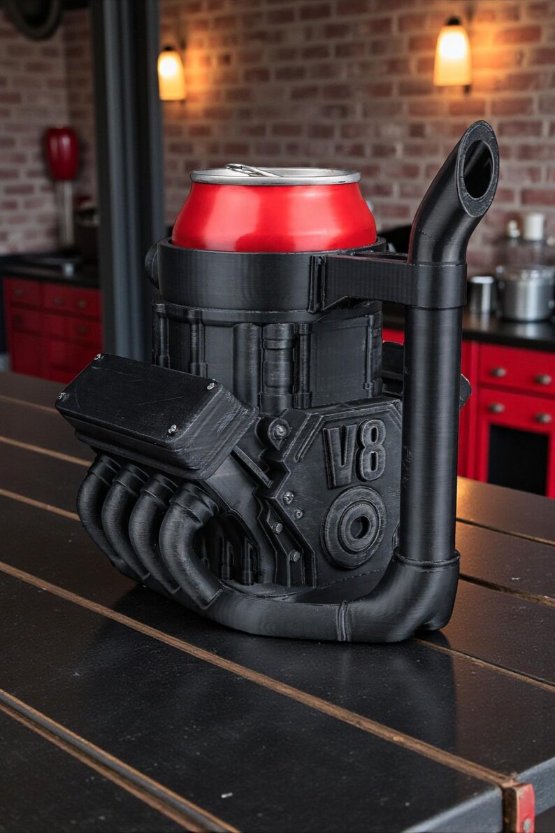 May include: A black V8 engine-shaped can cooler holds a red can. The cooler has detailed mechanical features, including exhaust pipes and the letters "V8". It sits on a dark wooden surface, with a brick wall in the background.