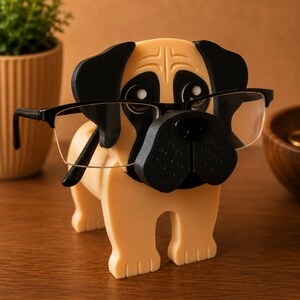 Pu&ograve; includere: Un portaocchiali a forma di carlino beige e nero. Il supporto ha un naso, orecchie e dettagli occhi neri. Gli occhiali hanno una montatura nera e poggiano sul naso del carlino. Il supporto &egrave; su una superficie di legno.