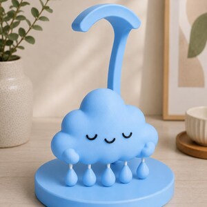 May include: A light blue cloud-shaped night light with a smiling face and dangling raindrop accents. The lamp has a curved handle and sits on a round base. The design is whimsical and suitable for a child's room.