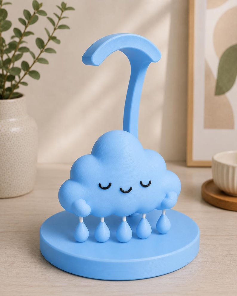 May include: A light blue cloud-shaped night light with a smiling face and dangling raindrop accents. The lamp has a curved handle and sits on a round base. The design is whimsical and suitable for a child's room.