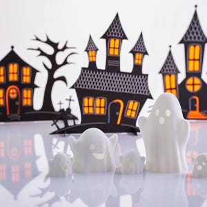 May include: Five white ghost figurines with smiling faces in front of a black paper backdrop of Halloween houses with orange windows.