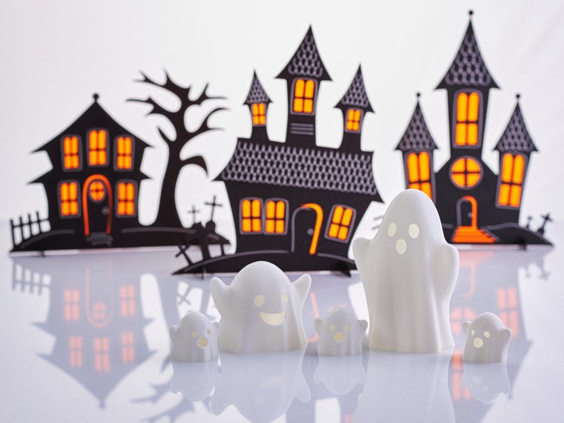 May include: Five white ghost figurines with smiling faces in front of a black paper backdrop of Halloween houses with orange windows.