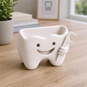 May include: A white, tooth-shaped container with a smiling face and arms holding a dental floss pick. The container has a rectangular opening at the top. The background includes a small potted plant, books, and a framed tooth illustration.