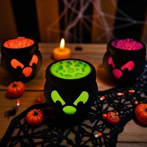 May include: Three black Halloween cauldrons with glowing faces and colorful contents: orange, green, and pink. Small pumpkins, a spider, and candy corn are scattered around the cauldrons on a wooden surface.