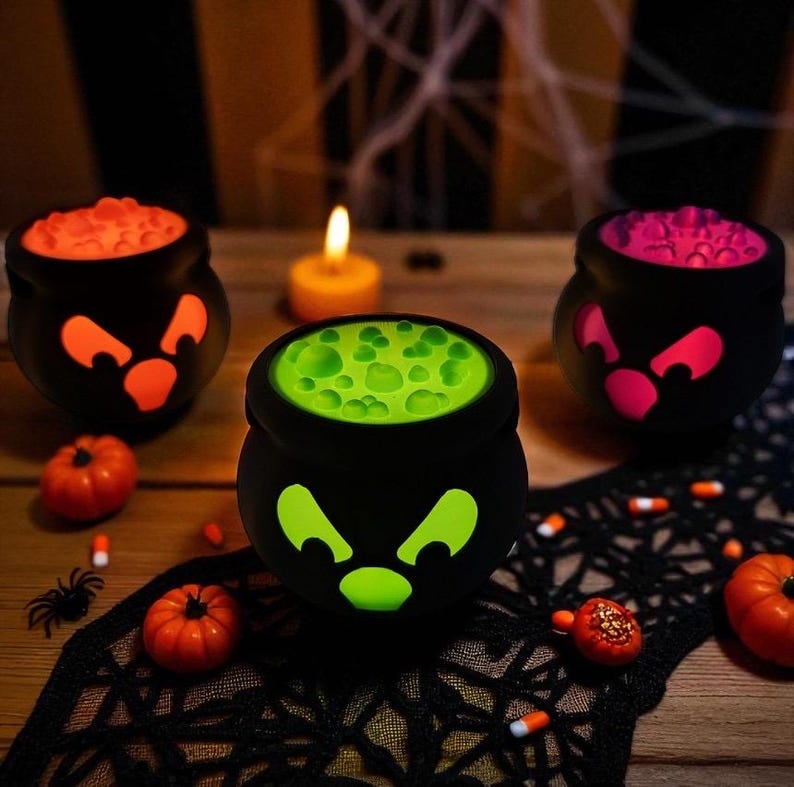 May include: Three black Halloween cauldrons with glowing faces and colorful contents: orange, green, and pink. Small pumpkins, a spider, and candy corn are scattered around the cauldrons on a wooden surface.