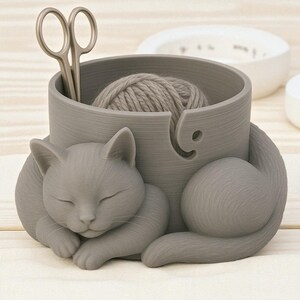 May include: A gray cat-shaped yarn bowl with a ball of yarn and silver scissors inside. The cat is curled up, appearing to be sleeping. The bowl has a slot for the yarn to feed through.