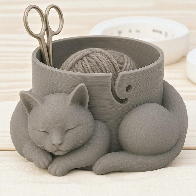 May include: A gray cat-shaped yarn bowl with a ball of yarn and silver scissors inside. The cat is curled up, appearing to be sleeping. The bowl has a slot for the yarn to feed through.