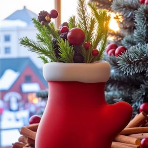 May include: A red mitten-shaped vase with a white rim, filled with green sprigs, red berries, and pinecones. The vase sits in front of a Christmas tree and a window with a blurred outdoor scene. Cinnamon sticks are in the foreground.