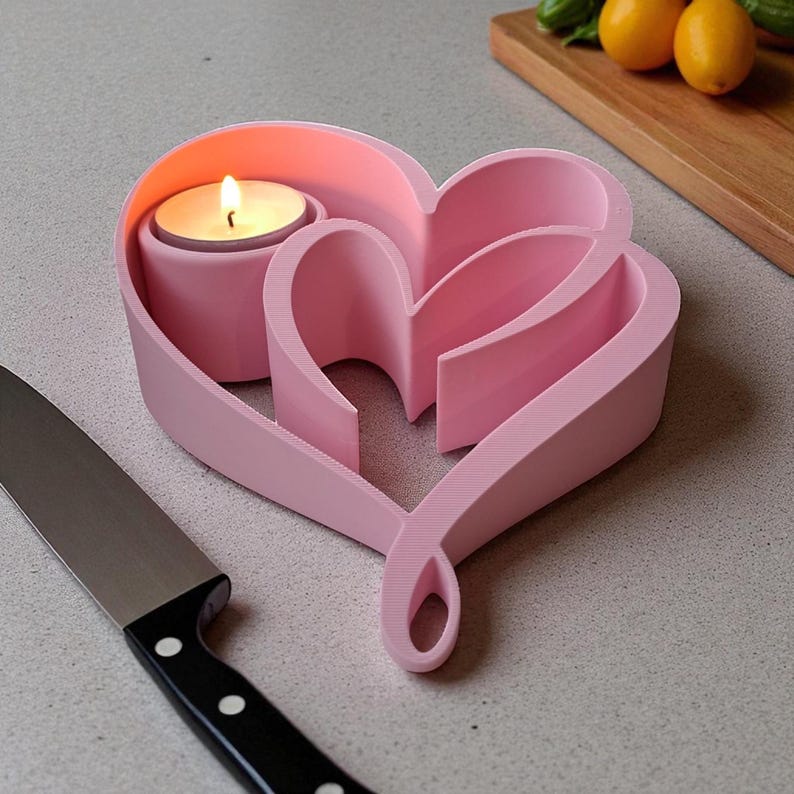 May include: A pink heart-shaped candle holder with a lit tea light. The holder has a layered design and a looped base. A black-handled knife is in the foreground, with a wooden cutting board and lemons in the background.