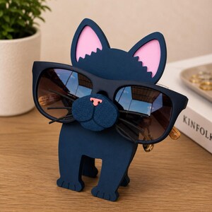May include: A navy blue cat-shaped eyeglass holder with pink inner ears and a textured blue nose. The holder is designed to hold a pair of sunglasses. The sunglasses have dark lenses and a gold-colored frame. The holder is on a wooden surface.