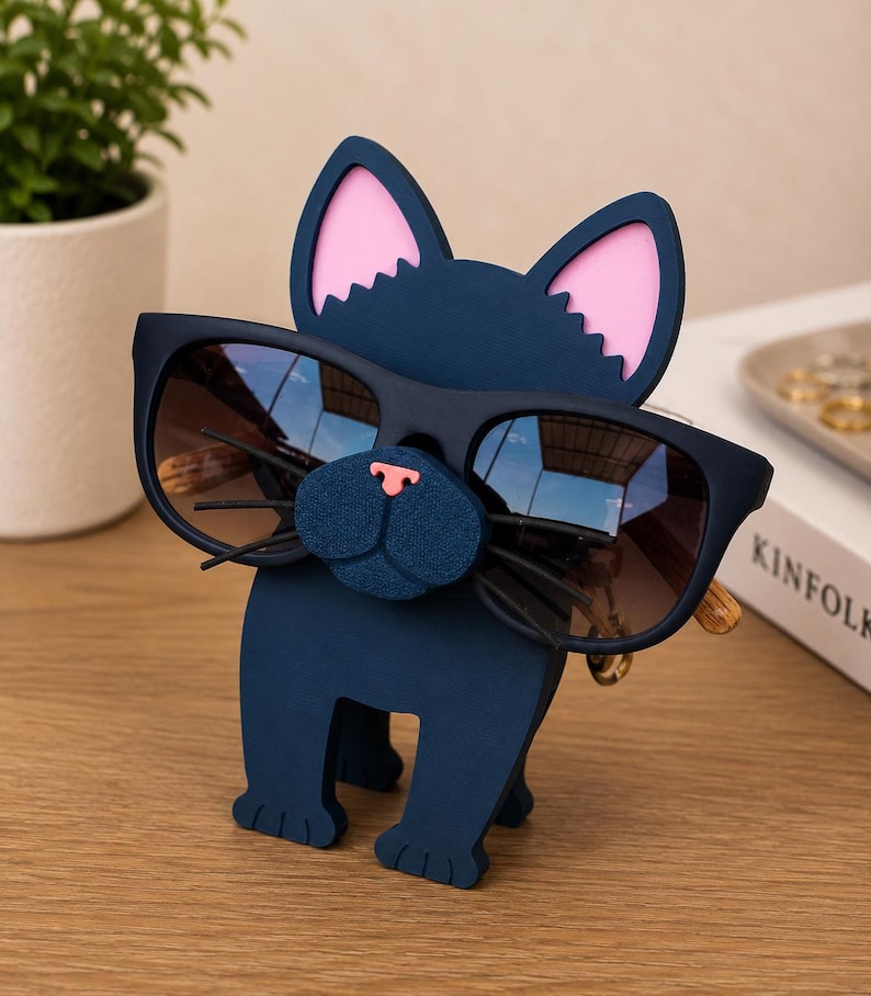 May include: A navy blue cat-shaped eyeglass holder with pink inner ears and a textured blue nose. The holder is designed to hold a pair of sunglasses. The sunglasses have dark lenses and a gold-colored frame. The holder is on a wooden surface.