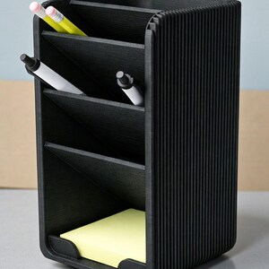 May include: A black desk organizer with multiple compartments. It holds pens, pencils, and a pad of yellow sticky notes. The organizer has a ribbed texture and a curved base, suitable for office or home use.