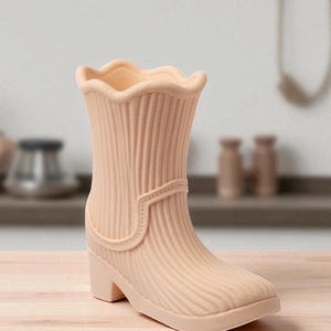 May include: A peach-colored boot-shaped vase with a ridged texture. The vase has a scalloped top edge, a strap detail, and a small heel. The design is reminiscent of a rain boot. The vase is made of ceramic or a similar material.