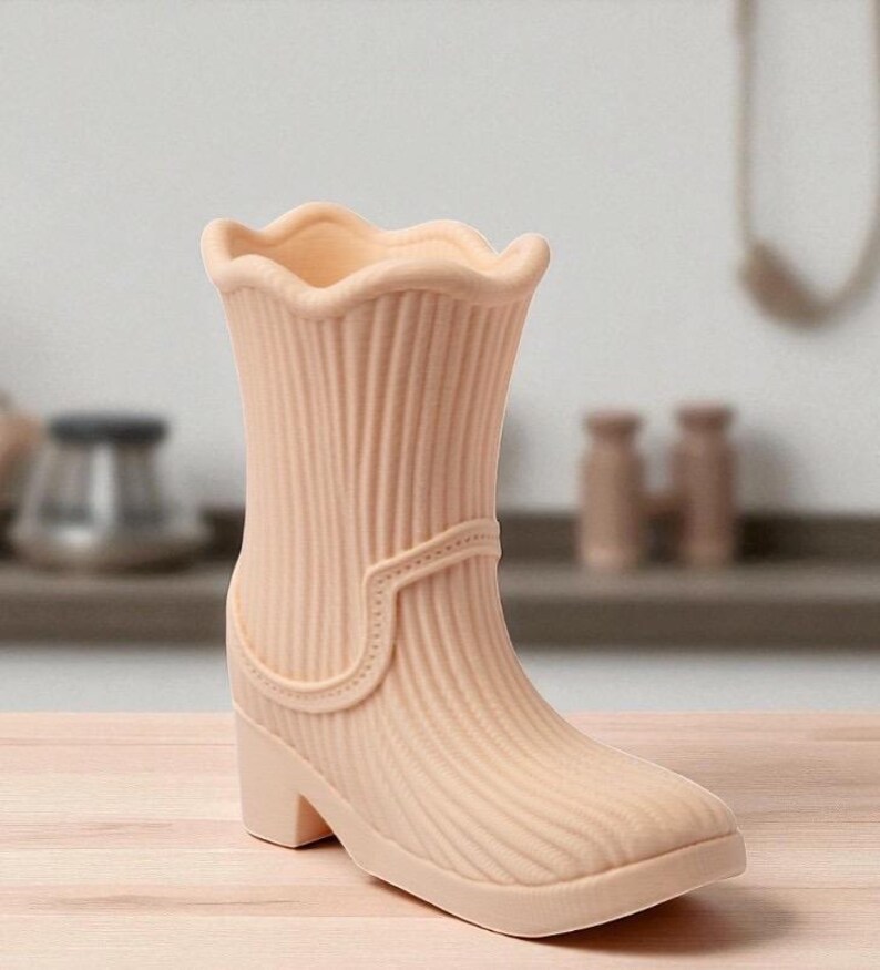 May include: A peach-colored boot-shaped vase with a ridged texture. The vase has a scalloped top edge, a strap detail, and a small heel. The design is reminiscent of a rain boot. The vase is made of ceramic or a similar material.
