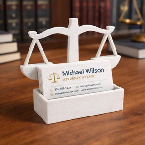 Puede incluir: Un tarjetero blanco con forma de balanza de la justicia. El soporte contiene tarjetas de visita con el nombre "Michael Wilson, Abogado". Las tarjetas muestran informaci&oacute;n de contacto, incluido un n&uacute;mero de tel&eacute;fono y una direcci&oacute;n de correo electr&oacute;nico.