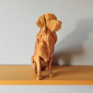 Op de afbeelding: Een goudkleurig hondenbeeld in zittende positie. De sculptuur heeft gedetailleerde vacht- en gelaatstrekken en wordt tentoongesteld op een houten plank. De hond kijkt naar rechts, met het hoofd iets gedraaid.