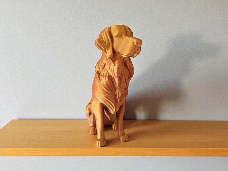 Op de afbeelding: Een goudkleurig hondenbeeld in zittende positie. De sculptuur heeft gedetailleerde vacht- en gelaatstrekken en wordt tentoongesteld op een houten plank. De hond kijkt naar rechts, met het hoofd iets gedraaid.