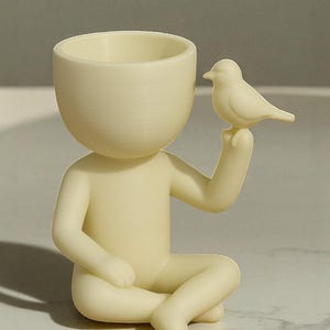 Op de afbeelding: Een cr&egrave;mekleurig beeldje van een zittend persoon met een kleine vogel. De figuur heeft een grote, open kop, ontworpen om een kleine plant in te houden. De vogel zit op de uitgestrekte hand van de figuur. Het beeldje is gemaakt van een glad, mat materiaal.