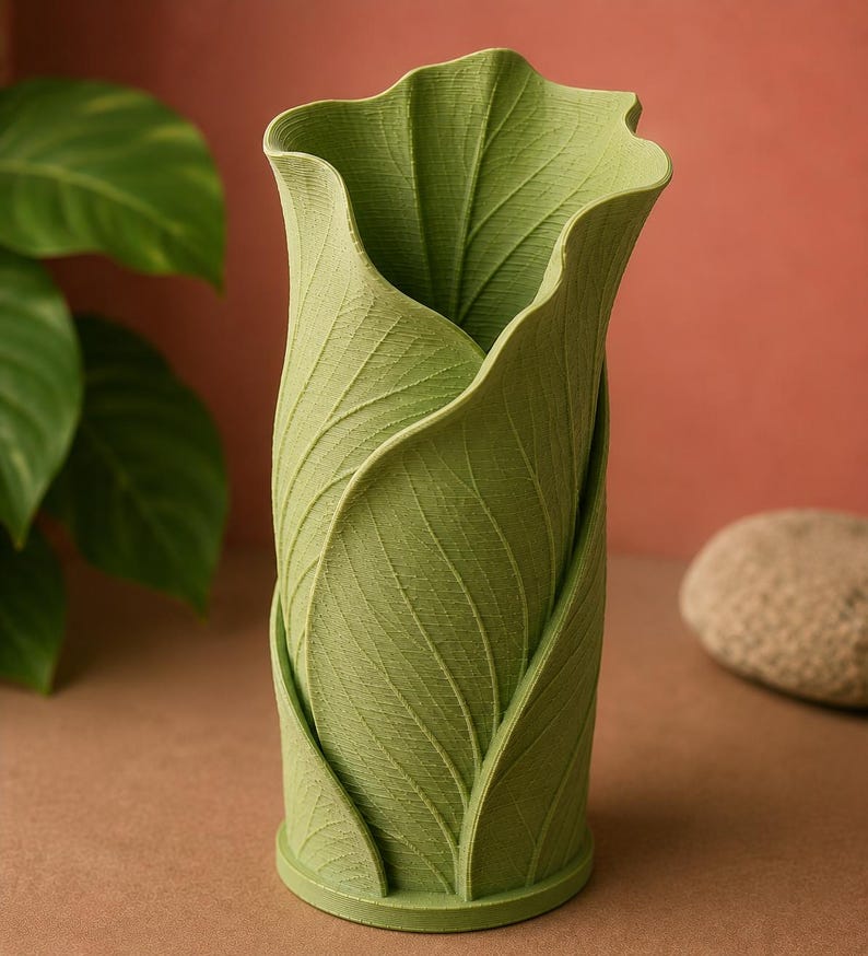May include: A green vase with a leaf design. The vase is cylindrical with a flared top, and the exterior is textured to resemble overlapping leaves. The vase is approximately 8 inches tall and has a round base.