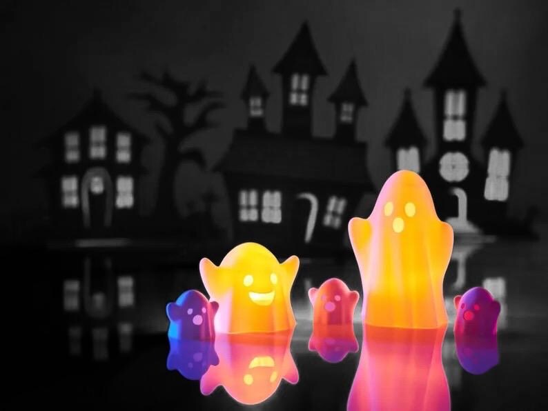 May include: A group of five glowing ghost-shaped Halloween decorations in front of a black and white silhouette of a haunted house. The ghosts are yellow, orange, and purple.