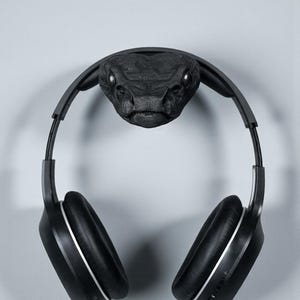 May include: A black headphone stand shaped like a snake head, with a pair of black headphones hanging from it. The snake head is textured and detailed, with visible eyes. The headphones have silver accents.
