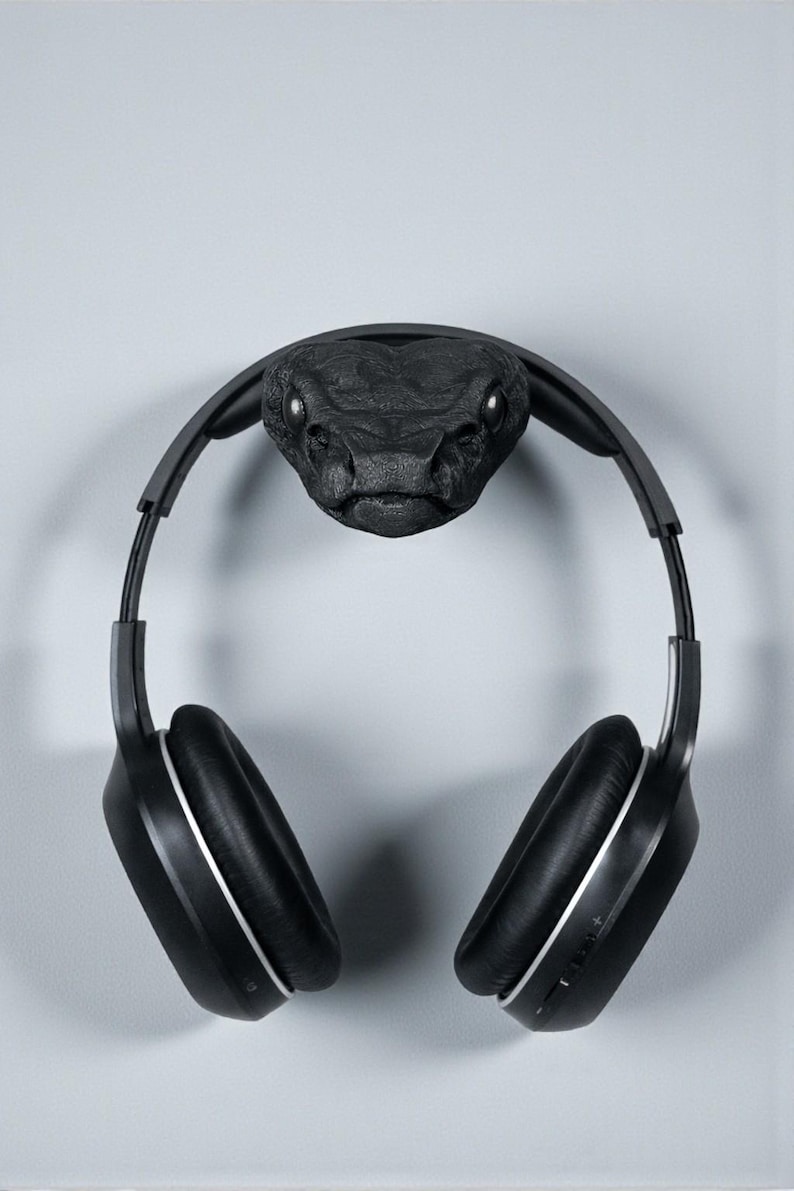 May include: A black headphone stand shaped like a snake head, with a pair of black headphones hanging from it. The snake head is textured and detailed, with visible eyes. The headphones have silver accents.