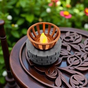 May include: A decorative candle holder with a copper-colored cage design, holding a flickering LED candle. The candle holder sits atop a gray, brick-patterned base. The candle holder is placed on a dark brown, ornate wooden table.