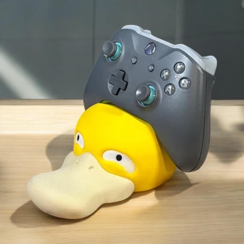 May include: A yellow Psyduck figure with a light-colored bill and eyes, supporting a gray Xbox controller. The controller has black buttons and teal accents. The figure is a unique gaming accessory.