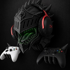 May include: A black dragon-themed wall mount helmet with a green interior, wearing black and red over-ear headphones. Two claw-shaped wall mounts hold a white wireless game controller on the left and a black wireless game controller on the right.