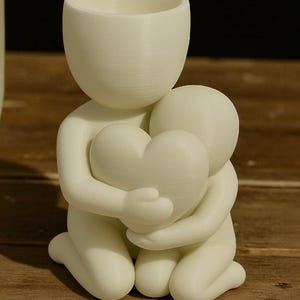 May include: A white figurine depicting two figures embracing a heart. The figures are kneeling, with one figure holding the heart. The figurine has a smooth, matte finish and is likely made of a 3D-printed material. The top figure has a cup-like shape.
