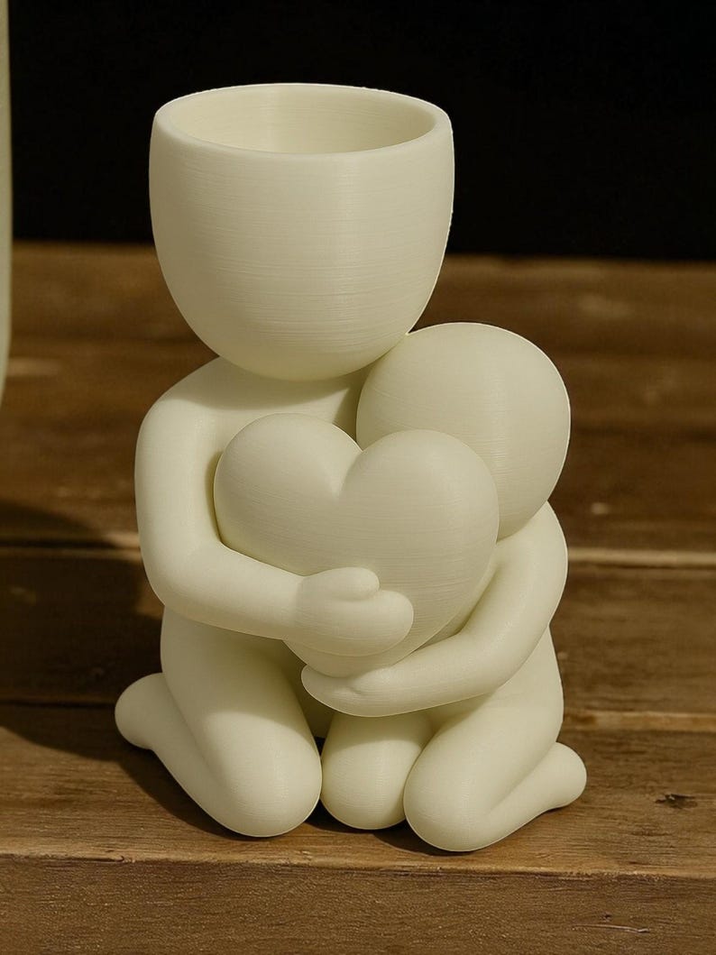 May include: A white figurine depicting two figures embracing a heart. The figures are kneeling, with one figure holding the heart. The figurine has a smooth, matte finish and is likely made of a 3D-printed material. The top figure has a cup-like shape.