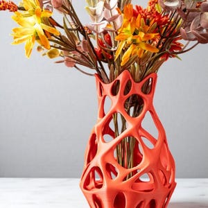 May include: An eye-catching orange vase with an open, lattice-like structure, filled with a colourful arrangement of artificial flowers. The flowers display shades of yellow, orange, and red, with green and brown foliage. The vase is a striking decorative piece.