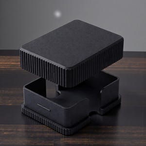 May include: A black, modern desk organizer with a rectangular top and base, and a ridged design on the sides. The top section is lifted, revealing an open compartment below. The organizer sits on a dark wooden surface.