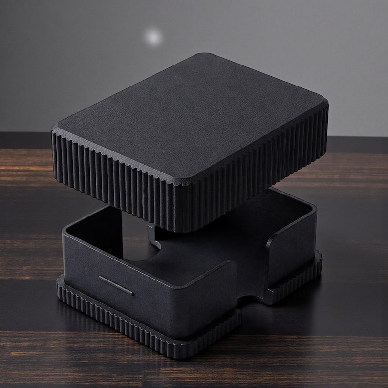 May include: A black, modern desk organizer with a rectangular top and base, and a ridged design on the sides. The top section is lifted, revealing an open compartment below. The organizer sits on a dark wooden surface.