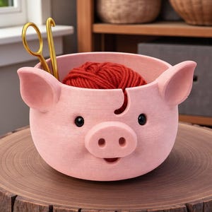Puede incluir: Un cuenco de hilo en forma de cerdo rosa con ojos negros y un hocico, que contiene una madeja de hilo rojo. Tijeras doradas y un ganchillo en su interior. El cuenco est&aacute; sobre una superficie de madera.
