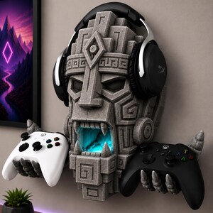 May include: A stone-textured wall mount shaped like a stylized ancient mask with a glowing blue interior. The mask features two stone hands extending from the sides to hold a white game controller on the left and a black game controller on the right.