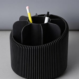 May include: A black desk organizer with a ribbed exterior and multiple compartments. A yellow pencil with a pink eraser and a white pen are inside the organizer. The organizer is designed to hold pens, pencils, and other office supplies.