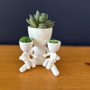 May include: A white ceramic planter shaped like a person, holding two smaller planters. The larger planter holds a succulent, while the smaller ones contain green plants. The design is minimalist and modern, suitable for small plants.