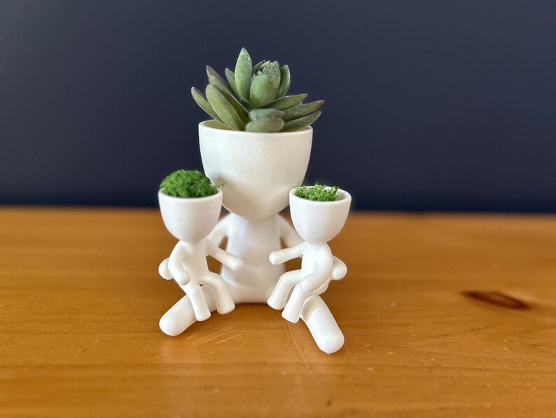 May include: A white ceramic planter shaped like a person, holding two smaller planters. The larger planter holds a succulent, while the smaller ones contain green plants. The design is minimalist and modern, suitable for small plants.