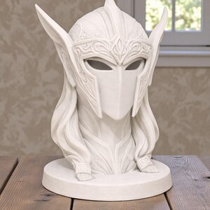 May include: A white sculpture of a helmeted figure with long hair and detailed facial features. The helmet has ornate patterns and pointed extensions. The sculpture sits on a round base, displayed on a wooden table.