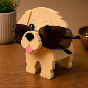 May include: A light tan dog-shaped eyeglass holder with black sunglasses. The dog has a black nose and a pink tongue. The sunglasses have a black frame and brown lenses. The holder is made of a soft material.