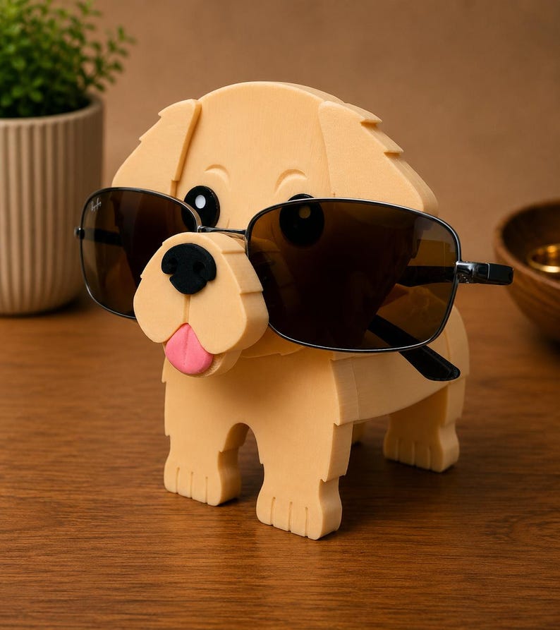May include: A light tan dog-shaped eyeglass holder with black sunglasses. The dog has a black nose and a pink tongue. The sunglasses have a black frame and brown lenses. The holder is made of a soft material.