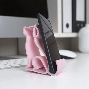 May include: A light pink phone stand shaped like a dog, holding a black smartphone. The stand has a curved design and is made of a smooth material. The phone is positioned vertically, displaying a dark screen. The stand is on a white surface.